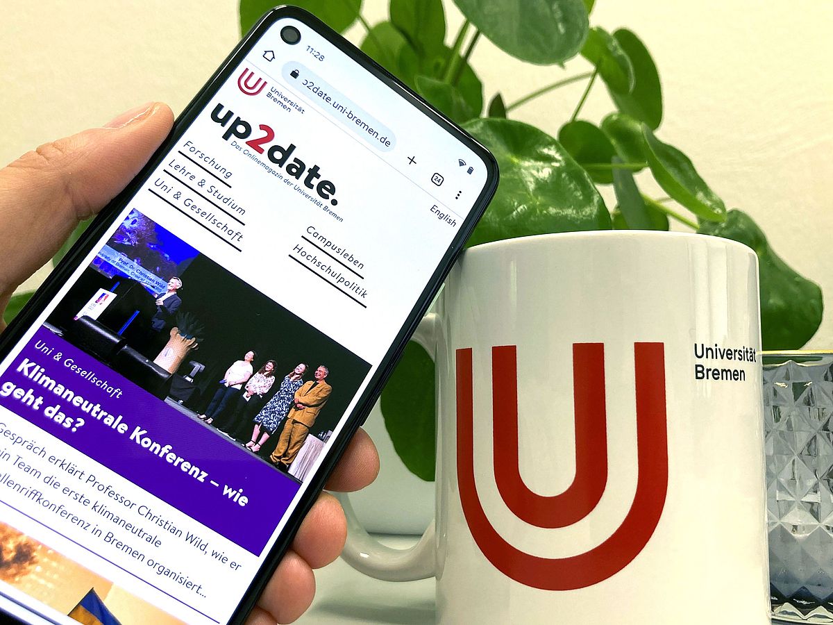 One hand holds a mobile phone on which the up2date. website is open. Next to it stands a coffee mug with the University of Bremen logo.