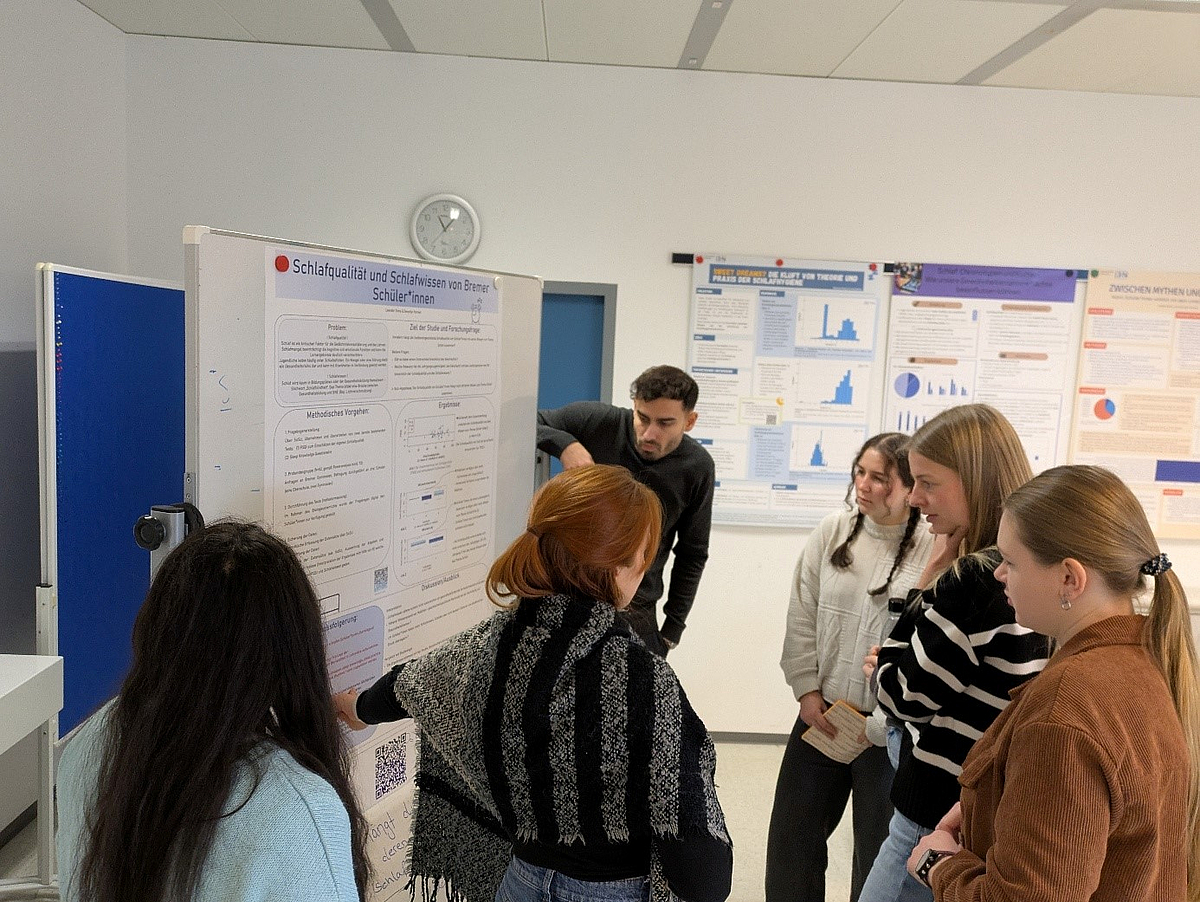 Gruppe von sechs Studierenden, die an einem wissenschaftlichen Poster steht und dieses betrachtet. Das Poster zeigt Ergebnisse eines Forschungsprojekts zum Thema Schlaf. Die Gruppe steht in einem Seminarraum mit weiteren Postern im Hintergrund.