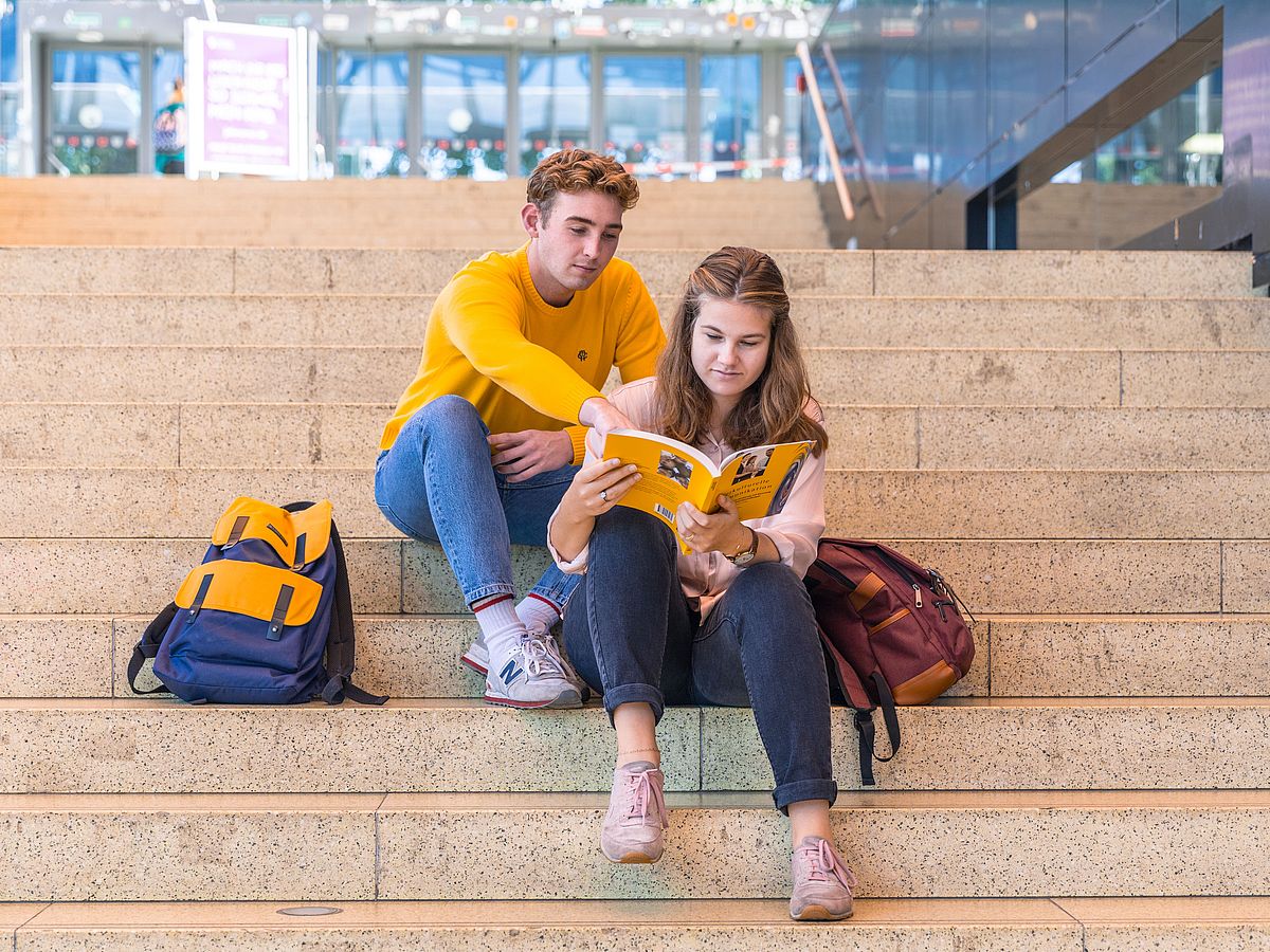 Zwei Studierende sitzen auf einer Treppe in der Glashalle.