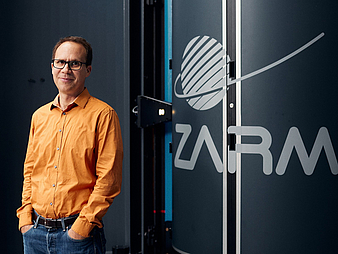 A man stands next to the ZARM logo and smiles into the camera. Marc Avila in front of the GraviTower Bremen Pro, where the gravitational forces on Mars can be simulated.