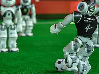 Roboter auf einem Spielfeld. Der rechte kickt den Ball.