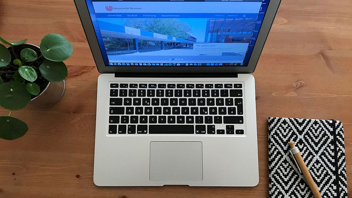 A laptop and a notebook next to a potted plant on a desk.